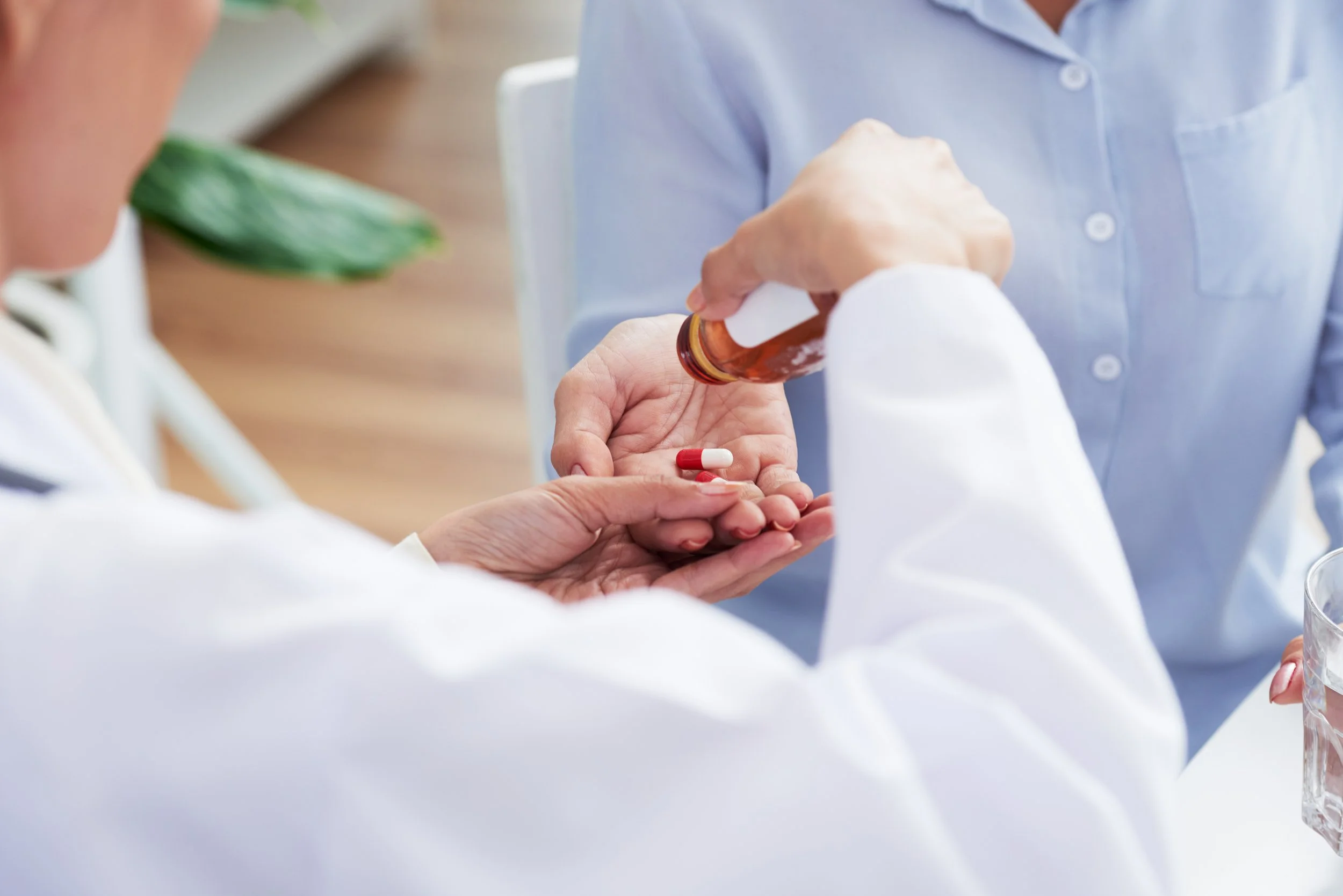 A person handing a red and white capsule pill to another person in a medical setting, with a pharmacy bottle. The person receiving the pill is wearing a white coat, and the background shows another individual in a light blue uniform.