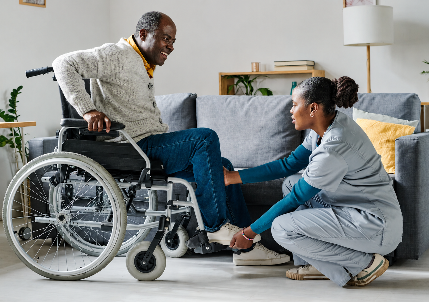 A healthcare professional assisting an elderly man in a wheelchair by adjusting his shoe laces or socks in a living room with a gray sofa, yellow pillows, and houseplants.