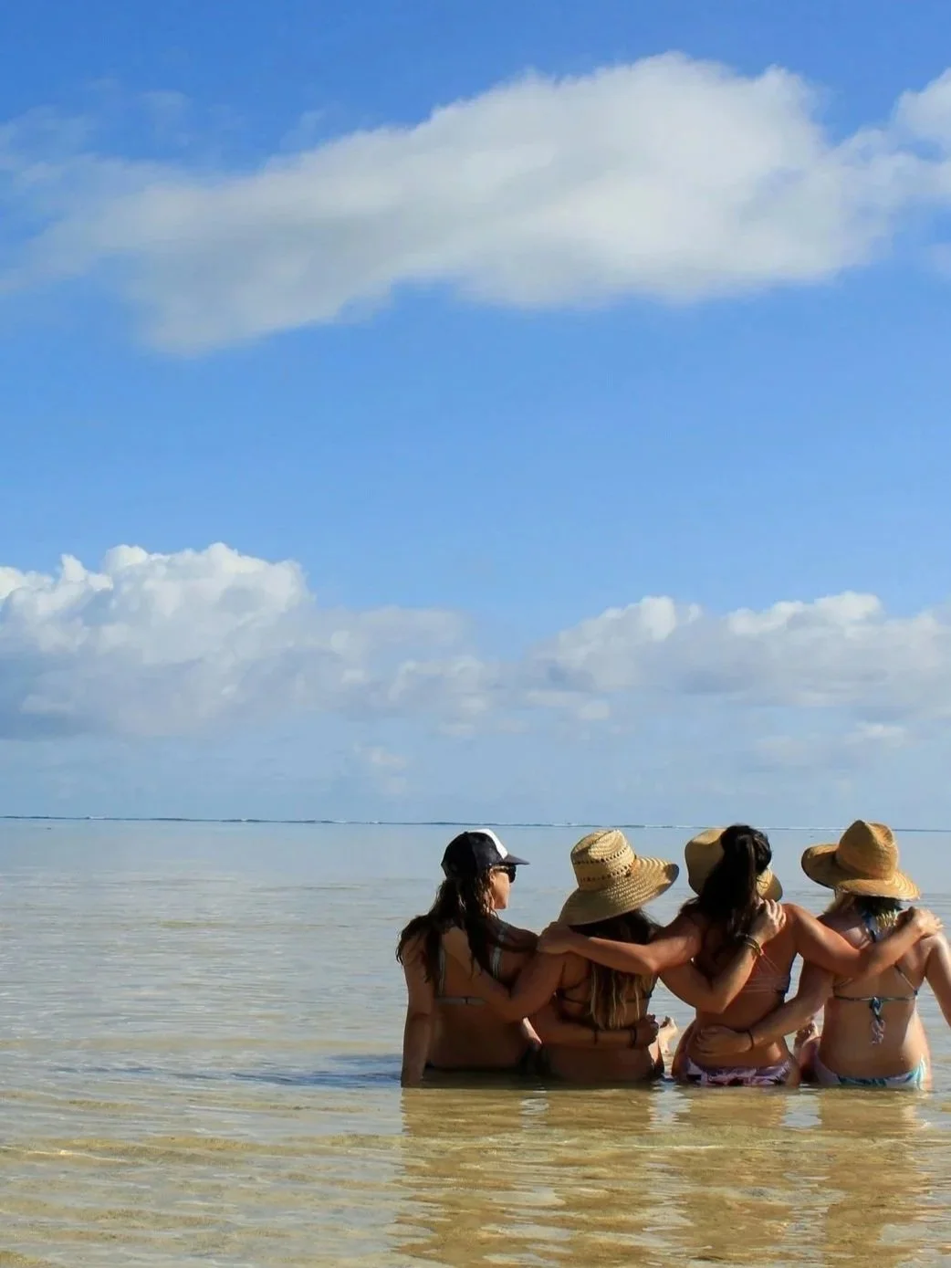 A group of women sitting together in shallow turquoise water on a sunny warm weather vacation