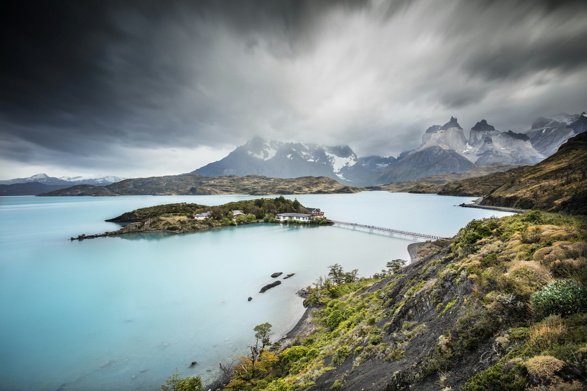 A mountain lake with turquoise water, surrounded by rocky slopes and green vegetation, under dark, cloudy skies.