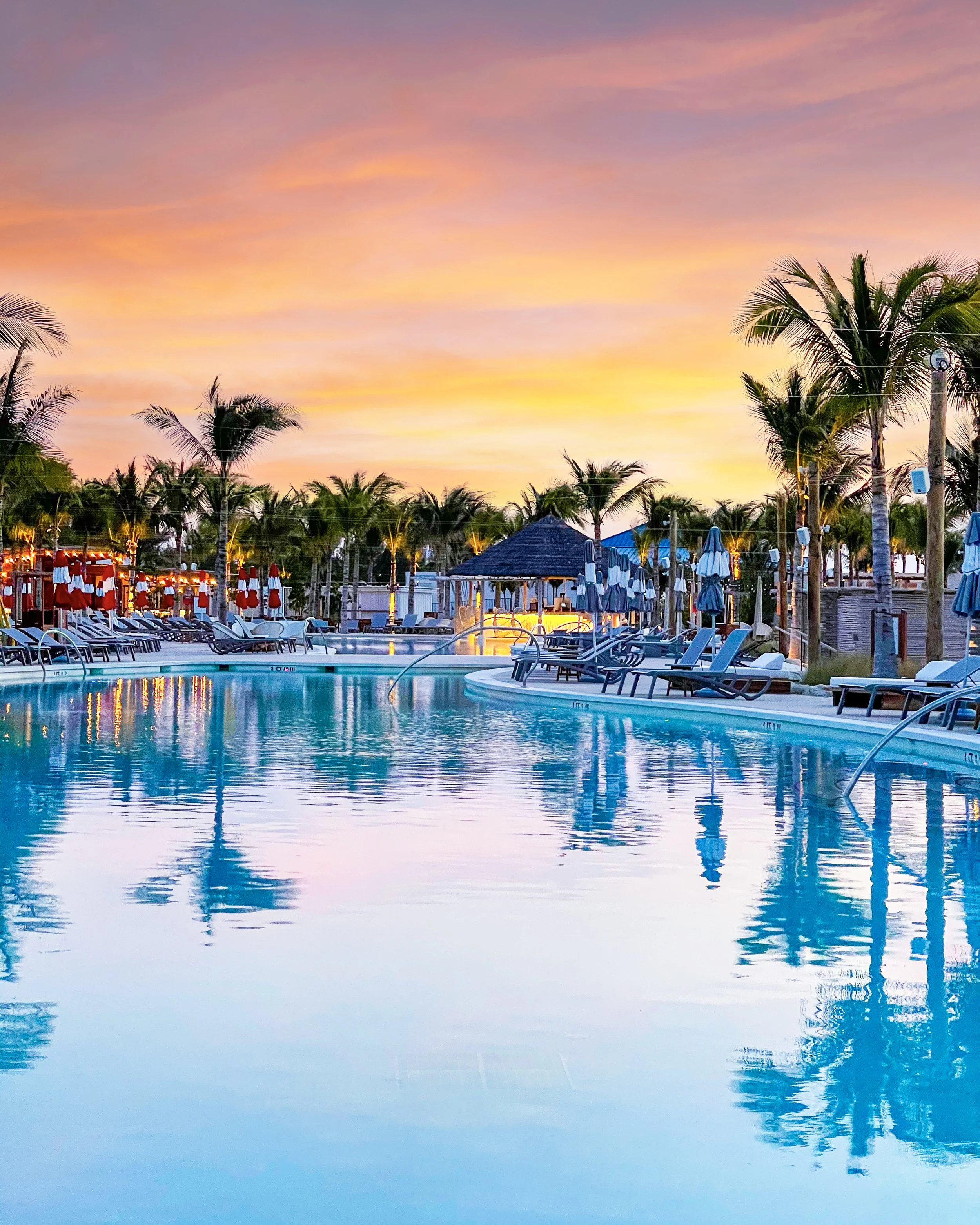 Sunset or sunrise sky above a tropical resort with palm trees, lounge chairs, umbrellas, and a swimming pool reflecting the colorful sky.