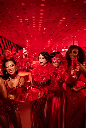 Four women dressed in red costumes celebrating in a festive environment, holding drinks and smiling.