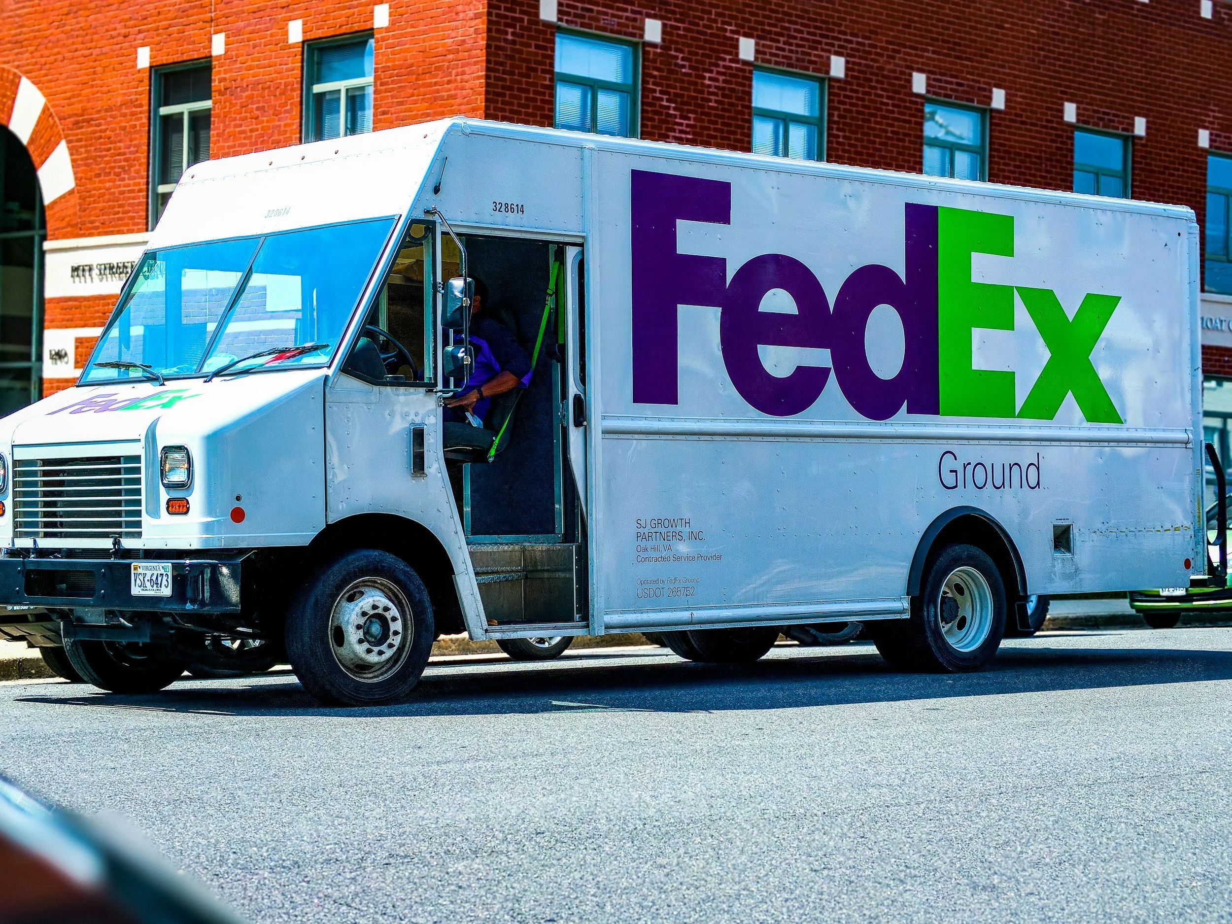 A FedEx delivery truck driving on a city street against a red brick building with multiple windows.