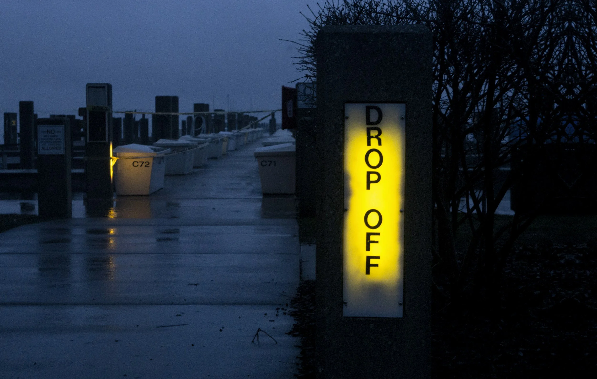A snowy marina at dusk with docked boats and a bright yellow illuminated sign that reads 'DROP OFF'.
