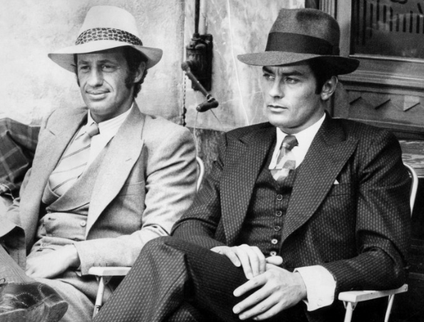Deux hommes en costume et chapeau assis, un homme souriant et l'autre sérieux, dans un décor intérieur.