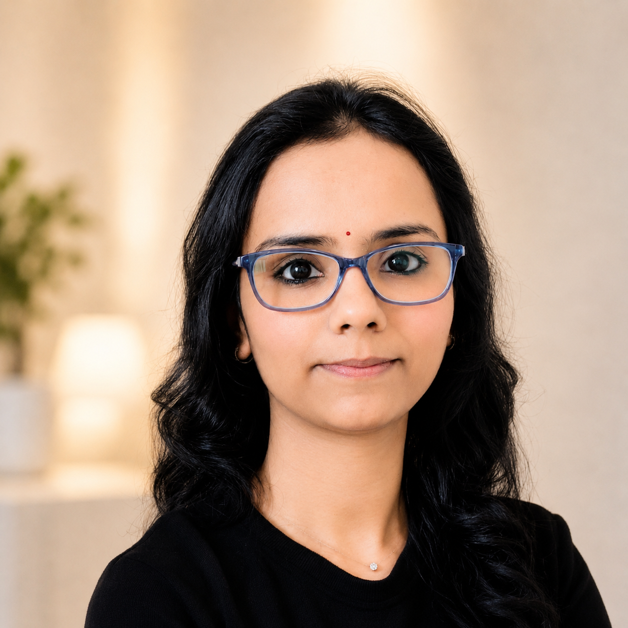 A woman with black hair, glasses, and a small red bindi on her forehead, wearing a black top, standing in a softly lit indoor setting.