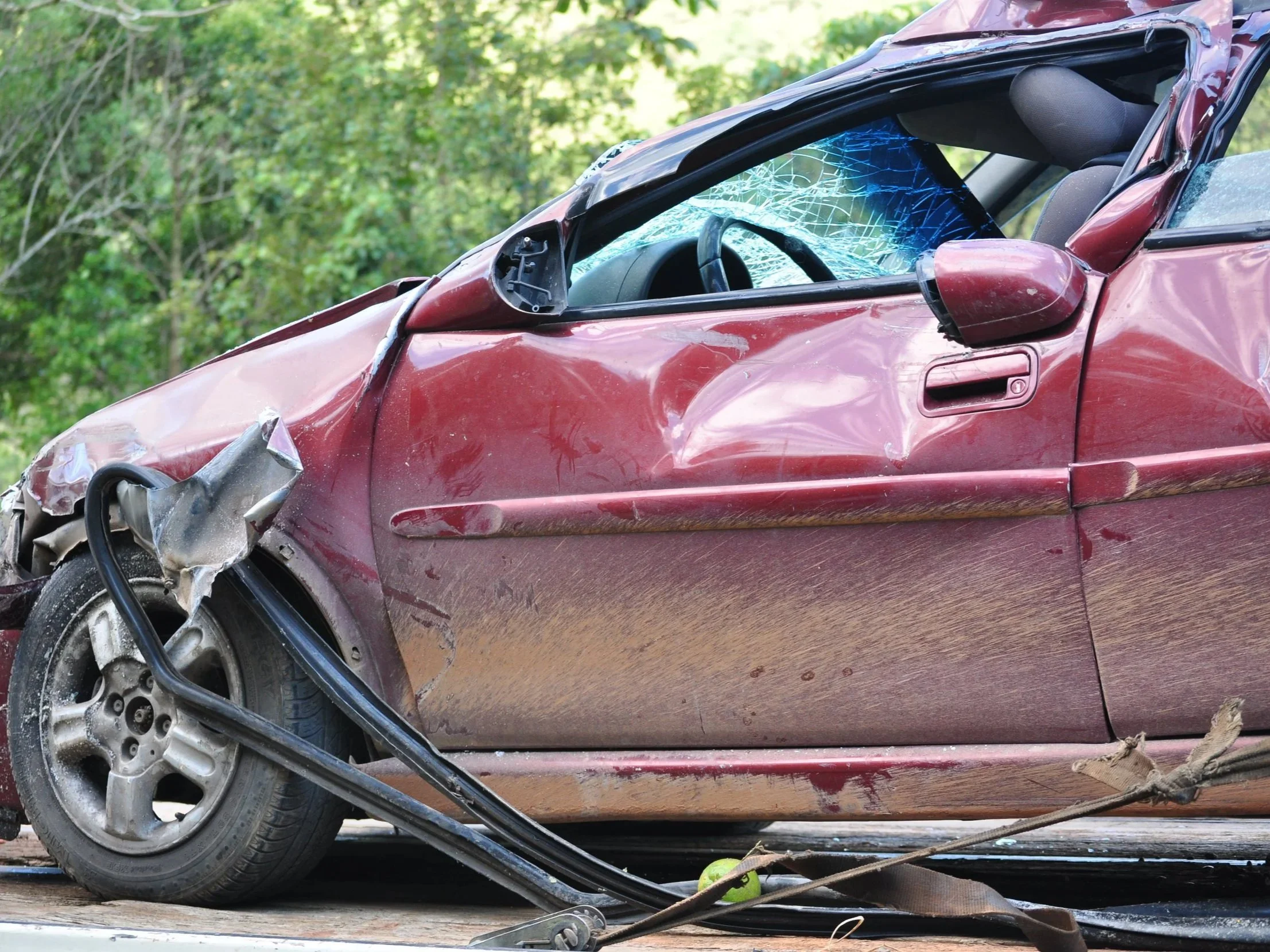 Red car with significant front-end damage, shattered windshield, and deployed airbags, parked outdoors with green foliage in background.