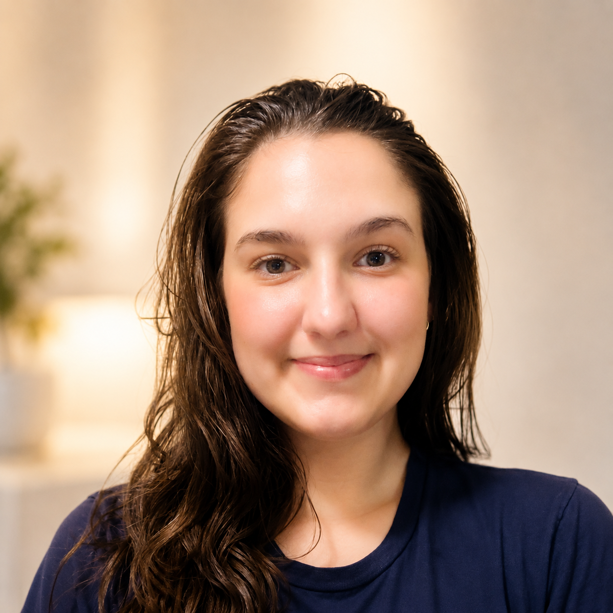 Close-up of a young woman with long, wavy brown hair, smiling softly, wearing a navy shirt, indoors with a blurred background.