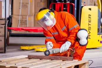 Worker in orange safety uniform and face shield using a hammer on a piece of wood in a warehouse.