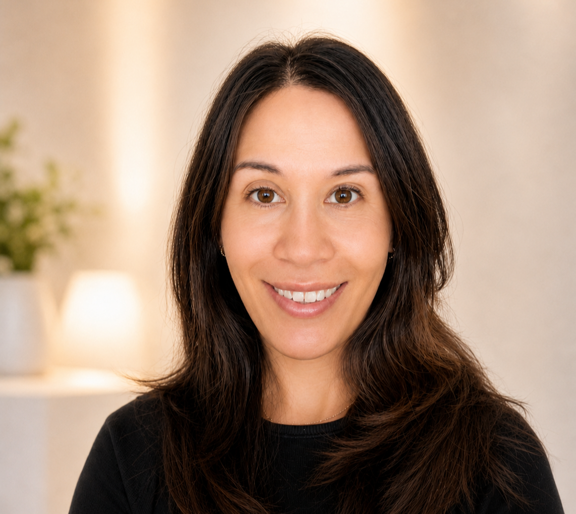 A woman with long brown hair smiling at the camera in a cozy indoor setting with a soft background featuring a lamp and a plant.