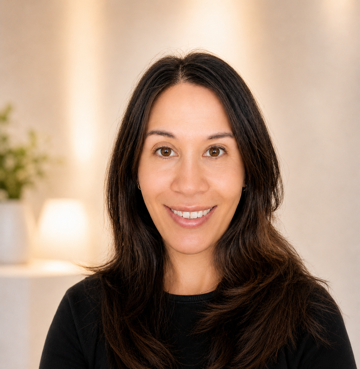 A woman with long, dark brown hair smiling indoors, with blurred background including a potted plant and a lamp.
