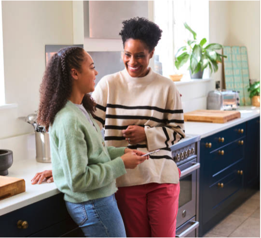 Two women smiling and talking in a bright kitchen, one holding a phone.
