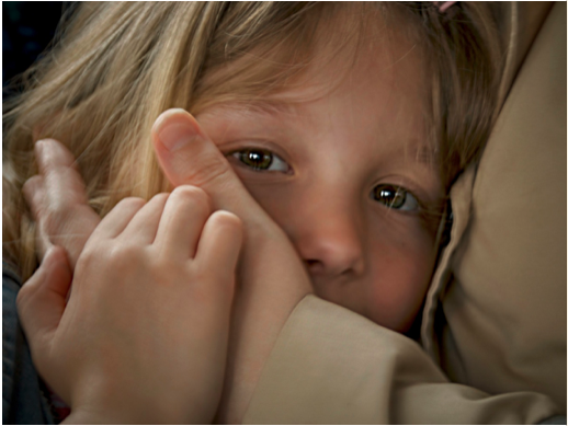 A young girl lying on a pillow with her hand covering part of her face, looking directly at the camera.