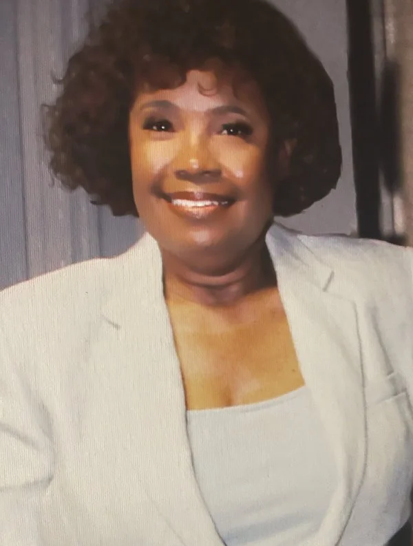A smiling woman with dark curly hair wearing a white blazer over a light top.