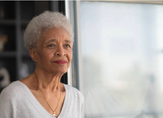 An elderly woman with gray hair looking thoughtfully out a window.