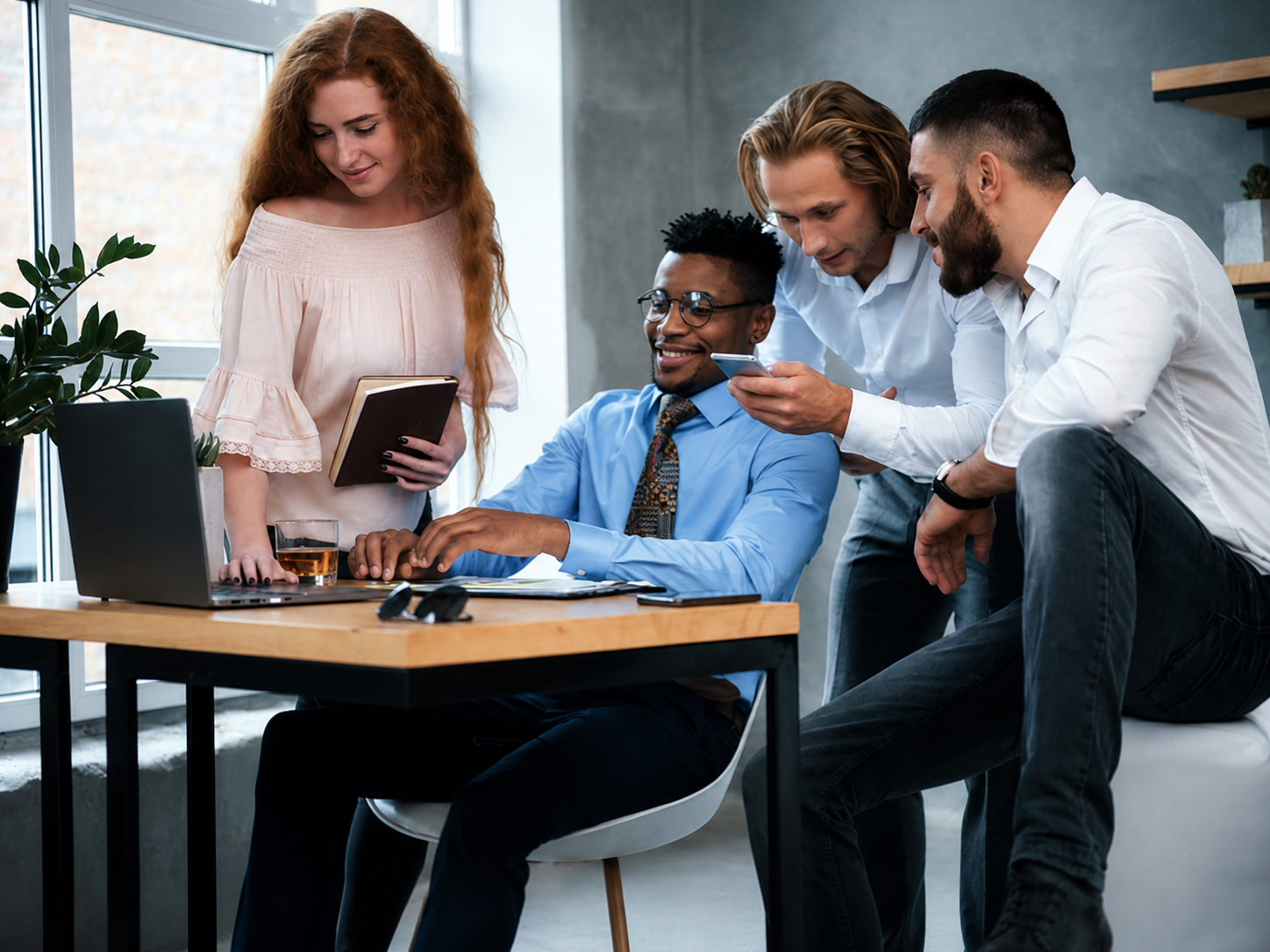 Four colleagues gathered around a laptop in a modern office, looking at a smartphone and discussing work.