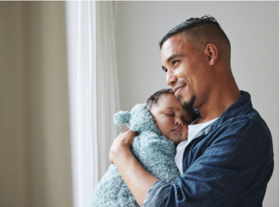 Man holding sleeping baby wrapped in blanket near window