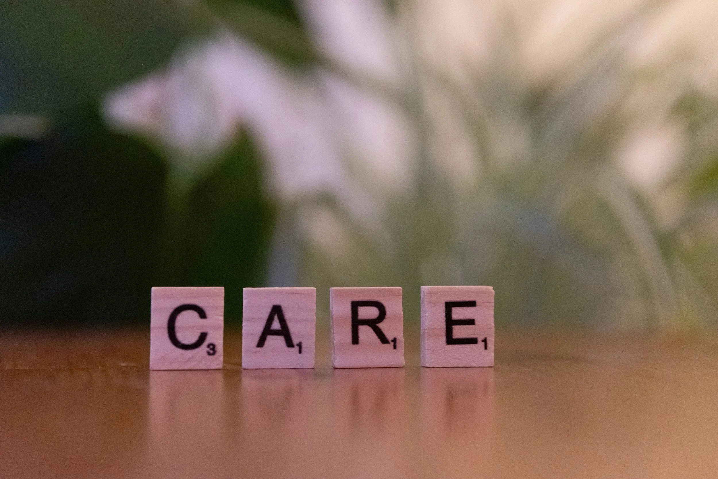 Close-up of Scrabble tiles spelling 'CARE' on a wooden surface, with blurred background including green leaves and white flowers.