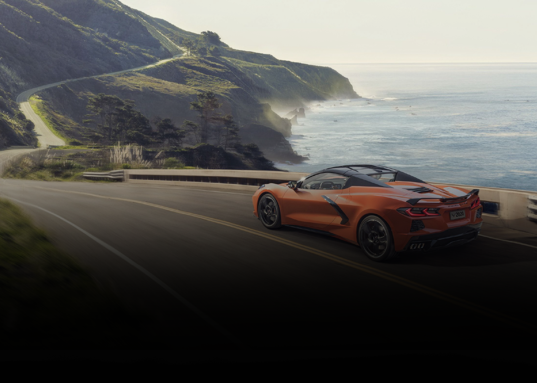 An orange sports car driving along a coastal mountain road with cliffs and ocean in the background.