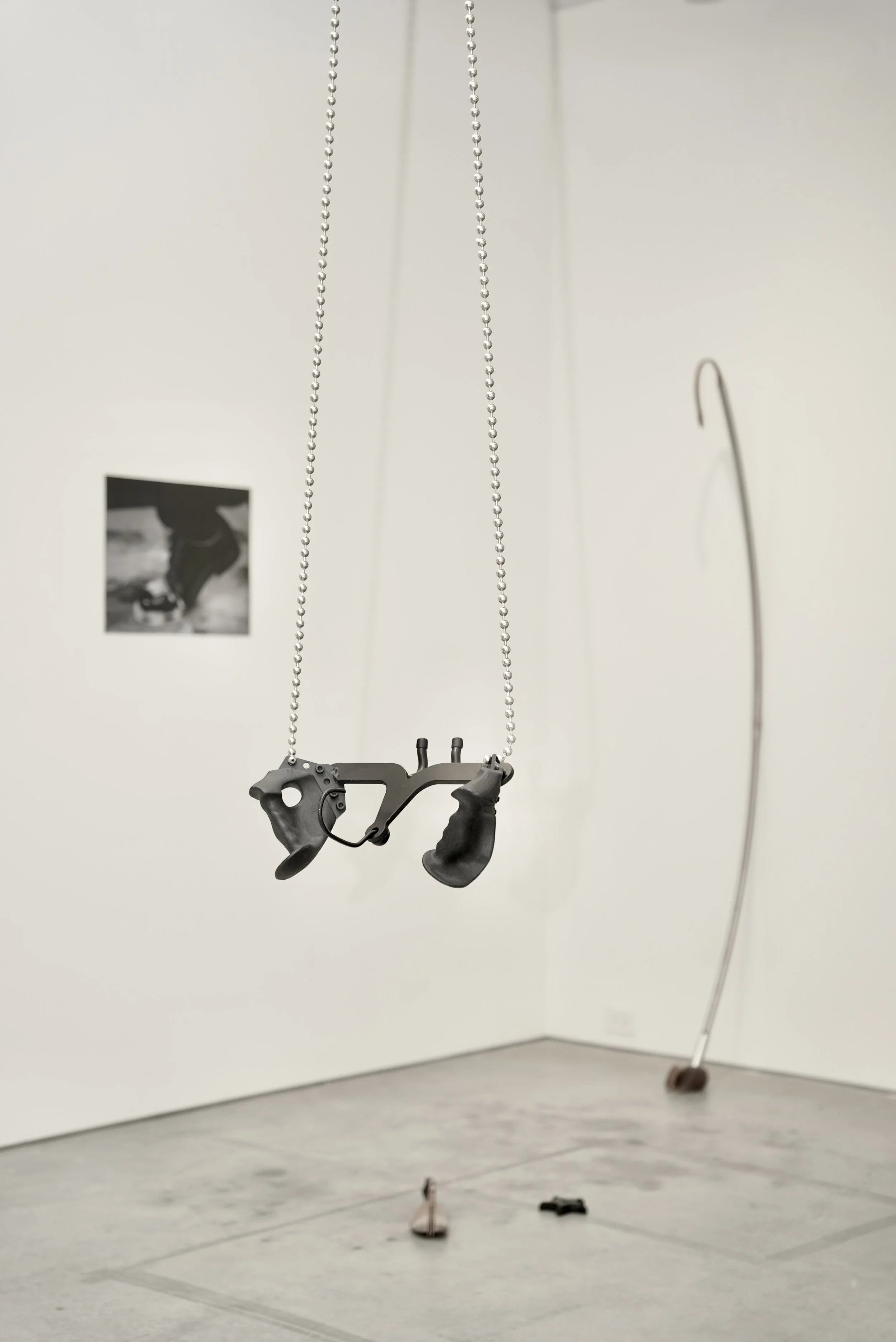 An art installation featuring a suspended black object resembling a medical or technological device, hanging from beaded chains in a gallery with white walls and gray floor. There is a small black and white photograph on the wall and a curved metal rod leaning against the wall.