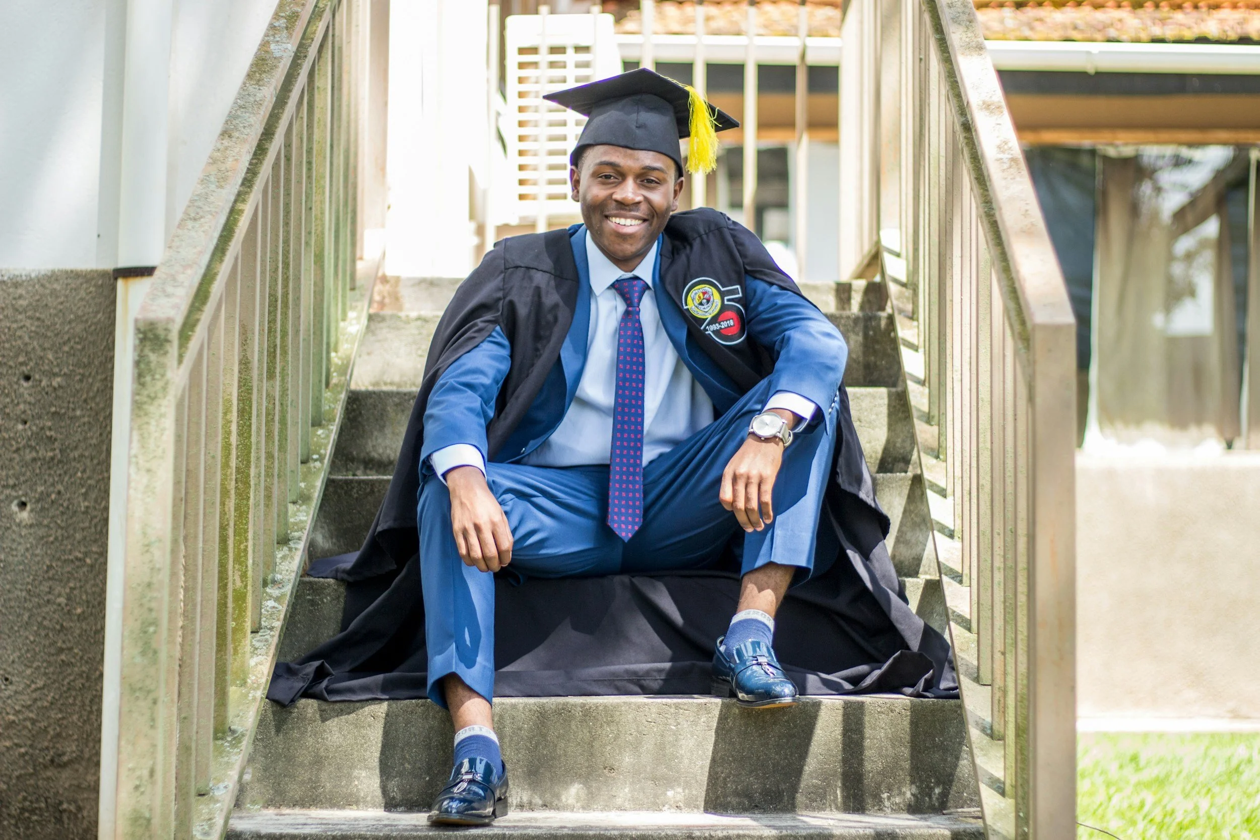 A young man in a graduation cap and gown sitting on steps outdoors, smiling, with a graduation patch on his gown.