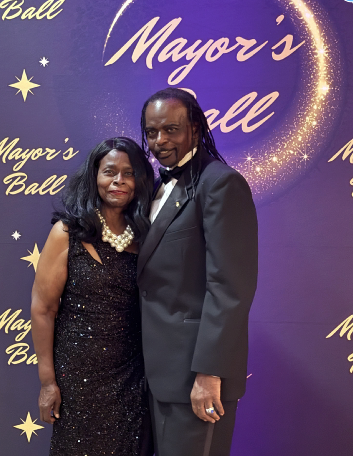 A woman in a sparkling black dress and a large pearl necklace standing next to a tall man in a black tuxedo with a bow tie, at a formal event called Mayor's Ball, with a purple backdrop decorated with gold stars and text.
