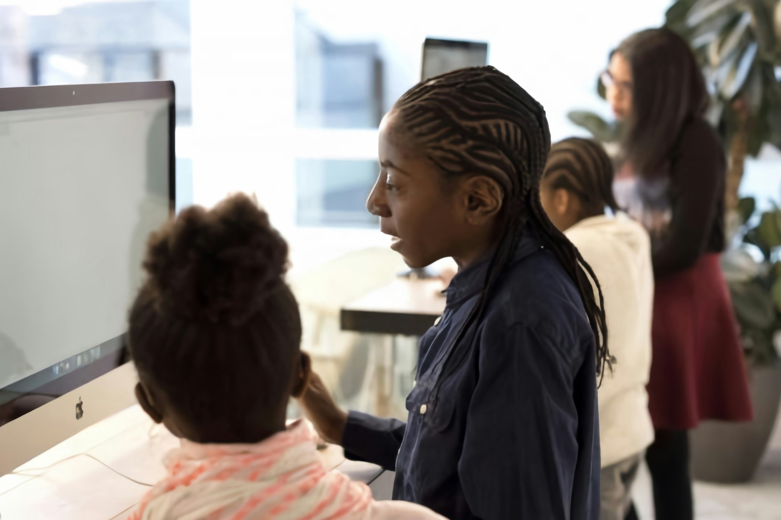 A diverse group of people working together in an office, with one woman with braided hair talking to a person sitting at a desk using an Apple computer.