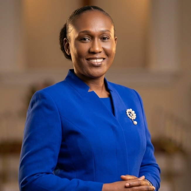 Professional portrait of a smiling woman wearing a royal blue suit, standing with folded hands inside a church sanctuary with softly blurred pews and warm lighting in the background.