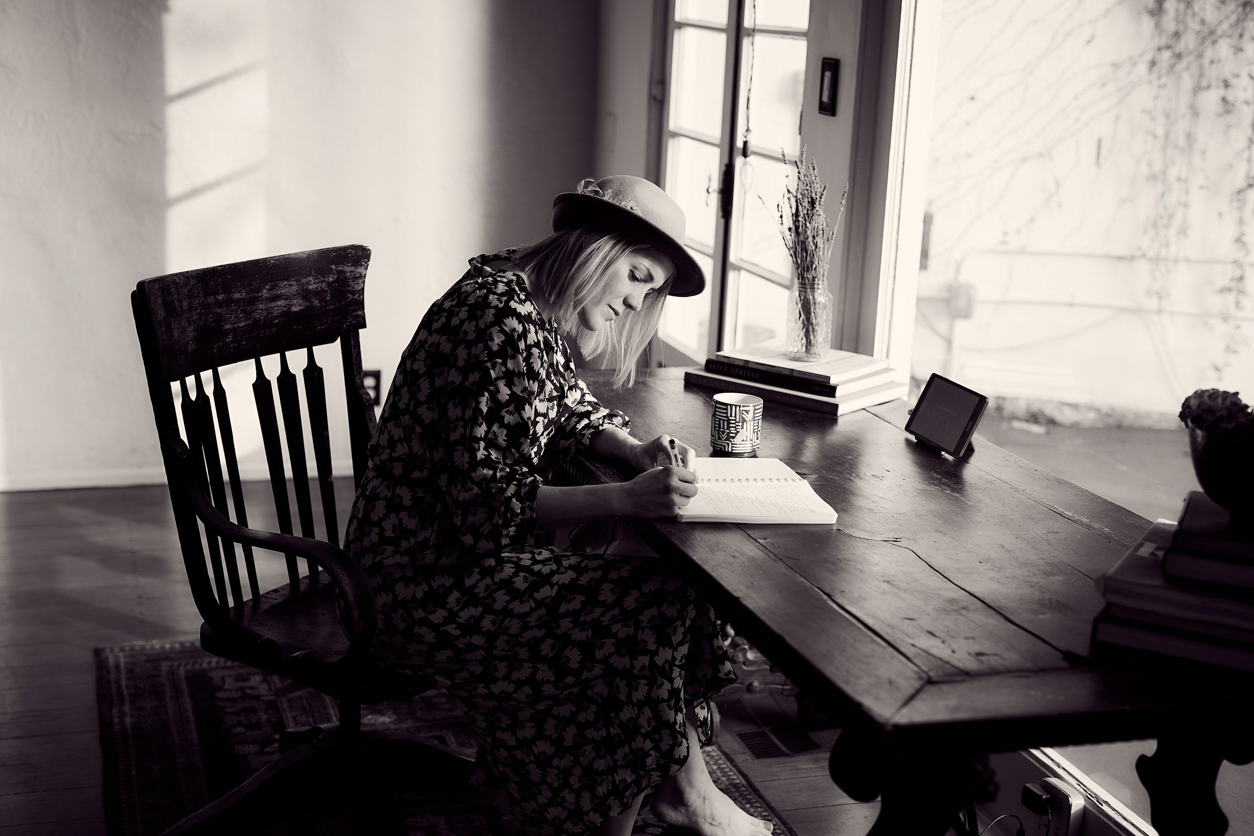 A woman sitting at a wooden desk writing in a notebook, wearing a hat and patterned dress, with sunlight coming through a window nearby.