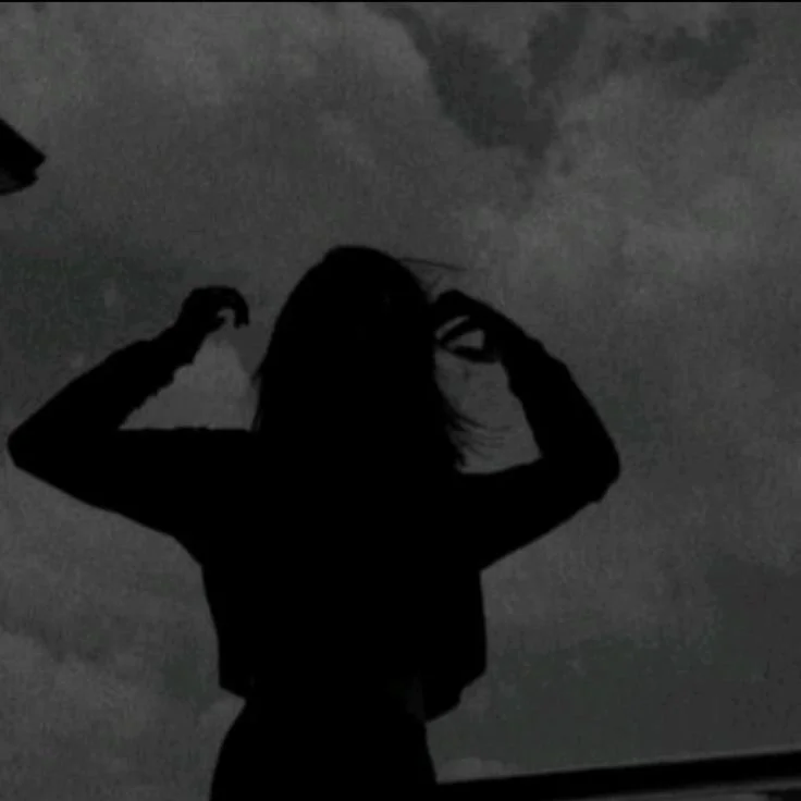 A grainy, low-contrast black and white photograph showing the dark silhouette of a person from behind. The person has long hair and is raising both hands toward their head as if fixing or running their fingers through their hair. The background consi