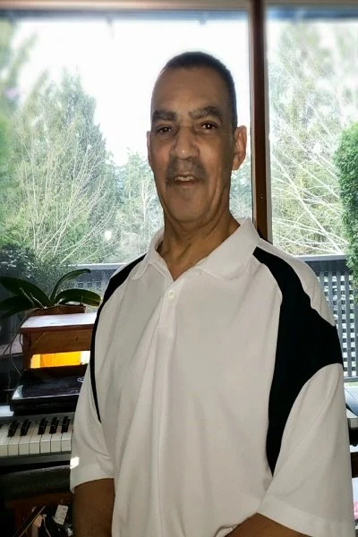 Portrait of a man wearing a white and black collared shirt, standing indoors in front of large windows with trees visible outside. A keyboard piano and houseplants can be seen in the background.