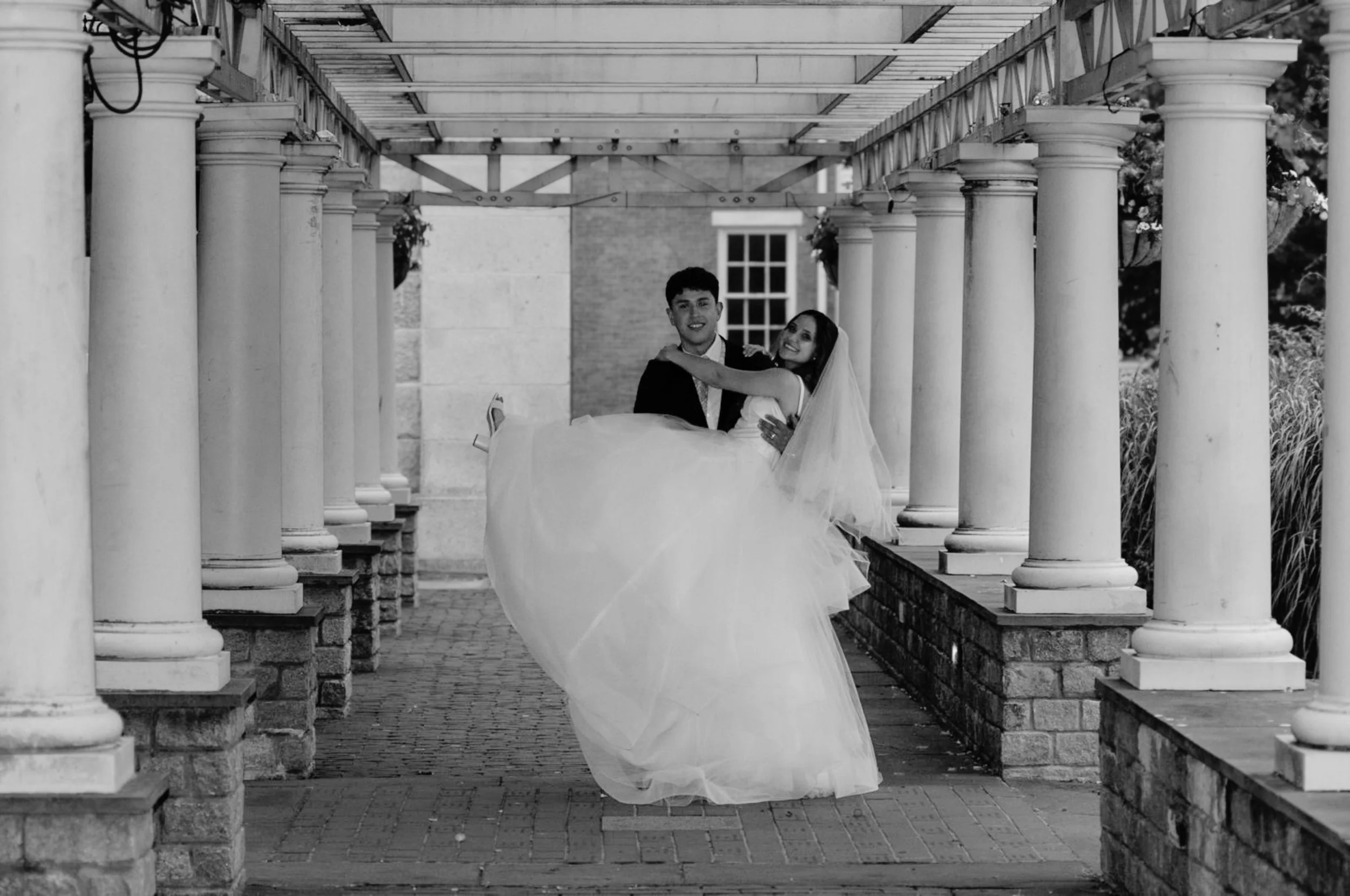 Black-and-white wedding photo of a groom carrying his bride beneath a columned walkway.