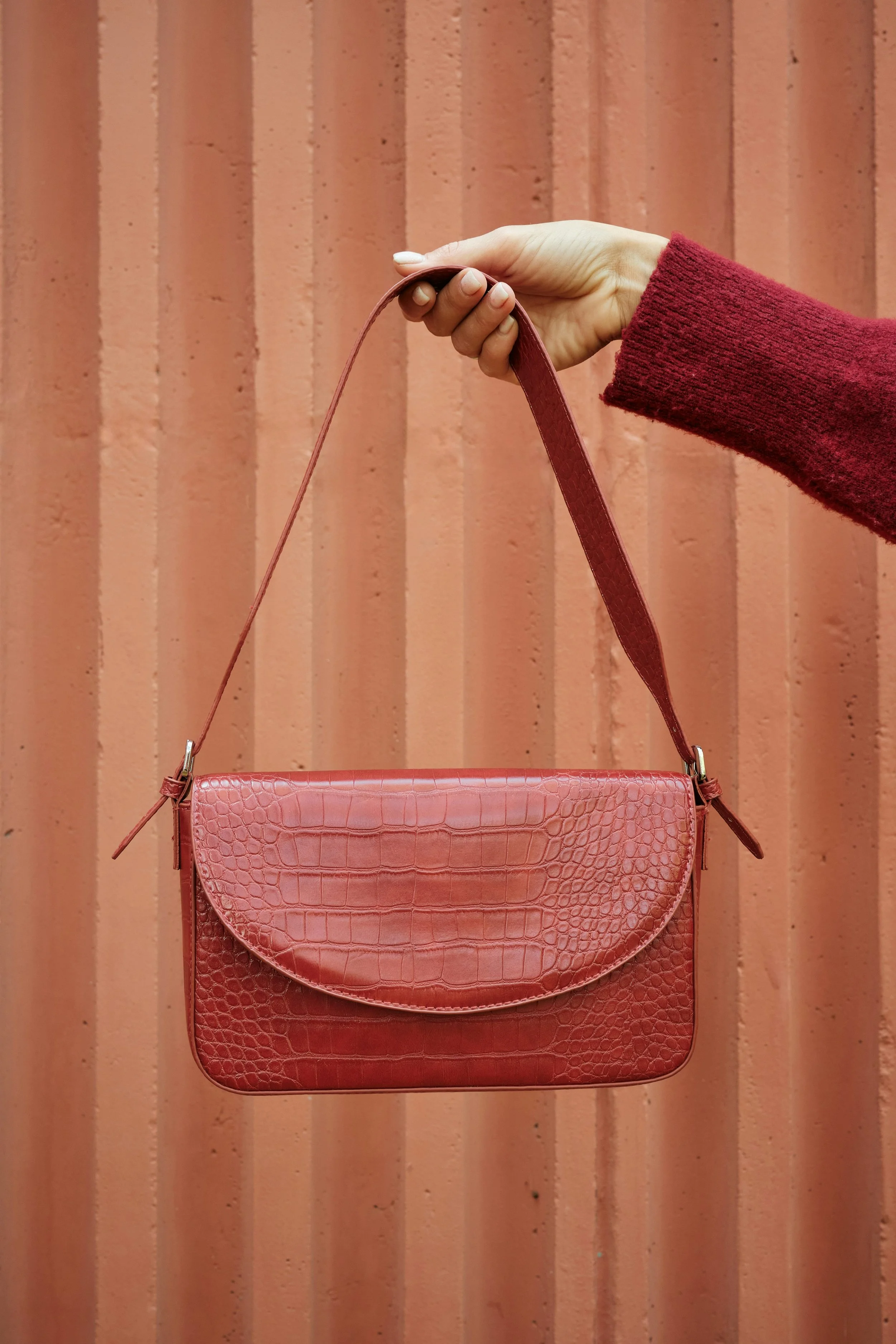 A person wearing a red sweater holding a pink crocodile-embossed handbag in front of a pinkish-orange wall with vertical lines.