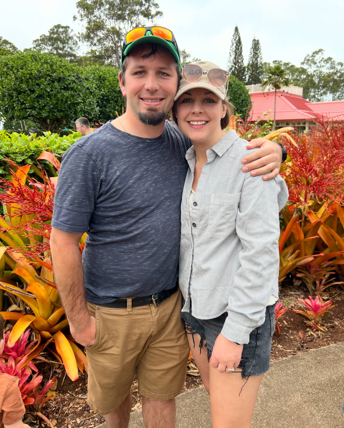 A smiling young man with a beard and a woman with a nose piercing and sunglasses on her hat standing outdoors in a garden. They are close together, with their arms around each other, smiling at the camera.