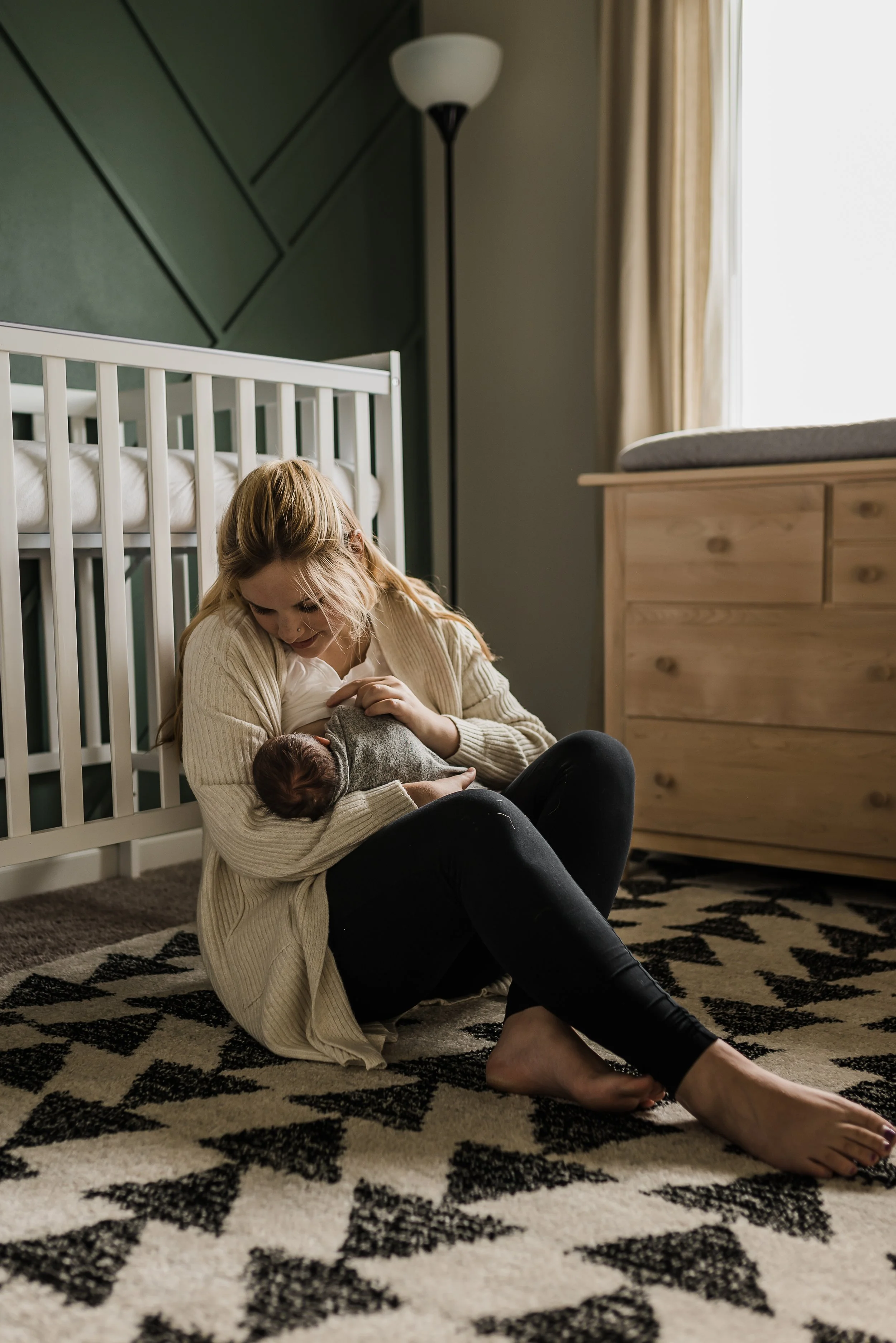 A woman with red hair and a beige sweater is sitting on a patterned rug, breastfeeding a baby in a gray onesie, inside a room with a crib, a wooden dresser, and a window with curtains.