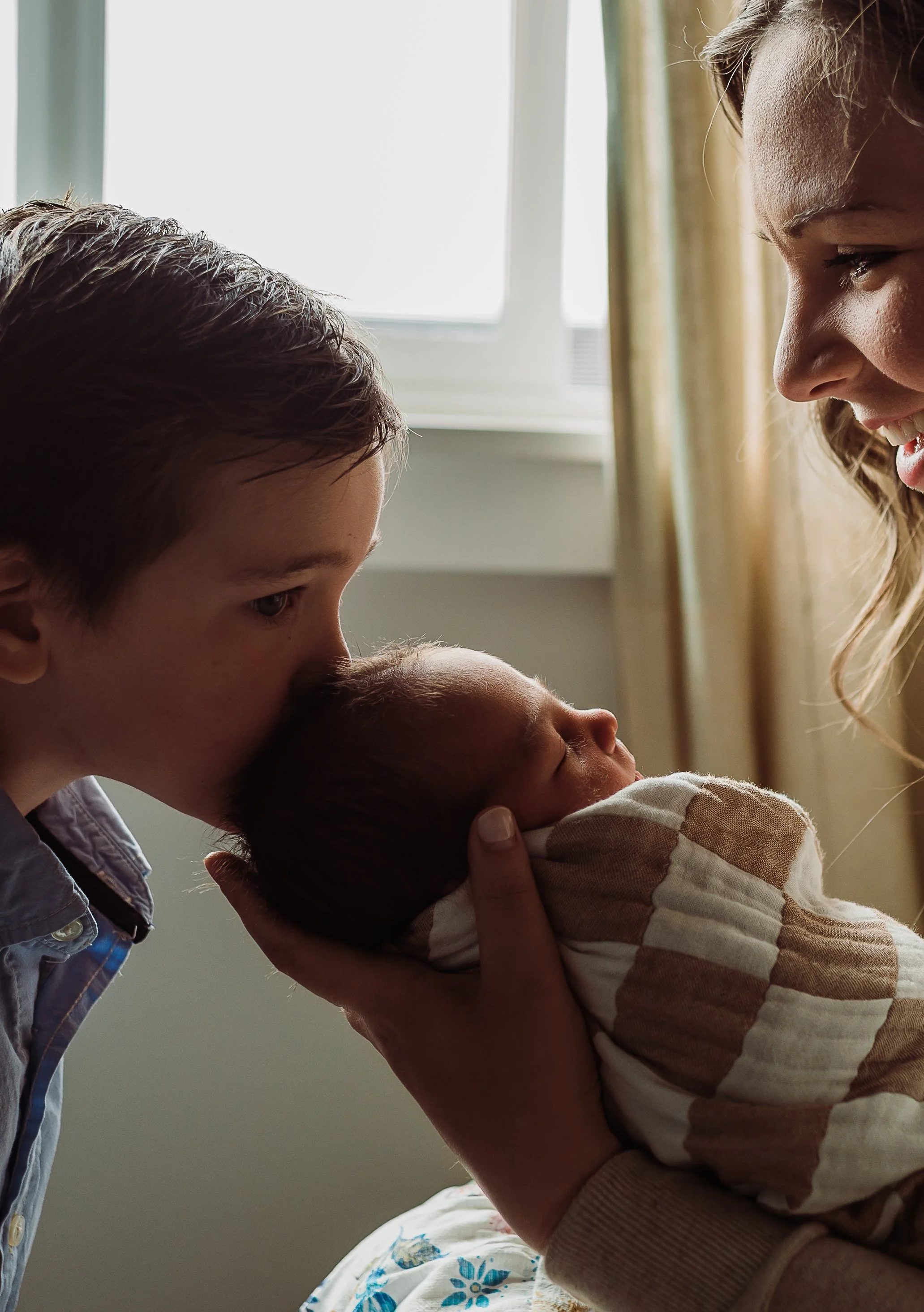 A woman is smiling and looking at an infant she is holding close, while a young boy leans in to look at the baby. They are indoors near a window with curtains.