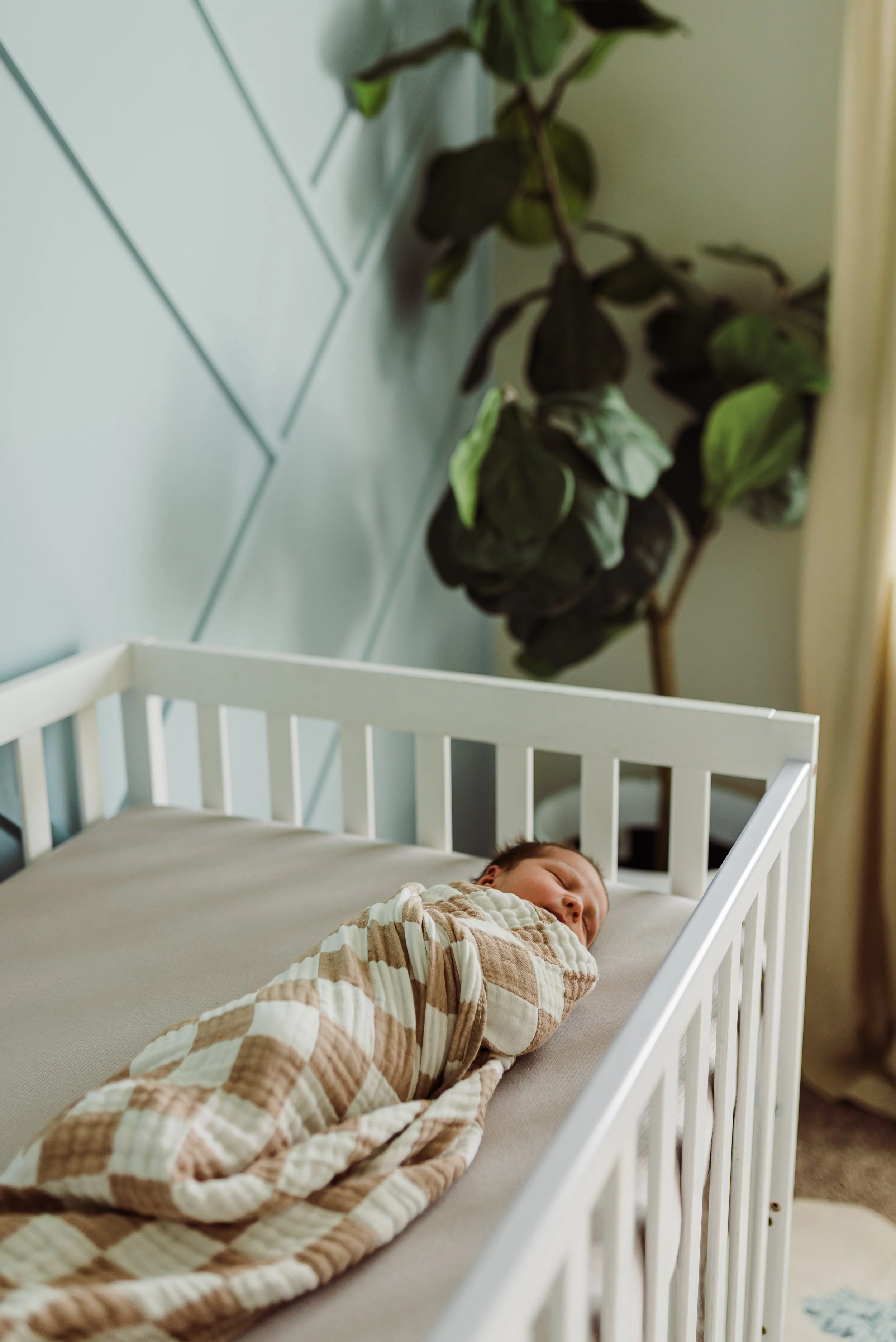 A baby sleeping in a white crib with a beige and white striped blanket, near a green plant on a wall with geometric pattern.