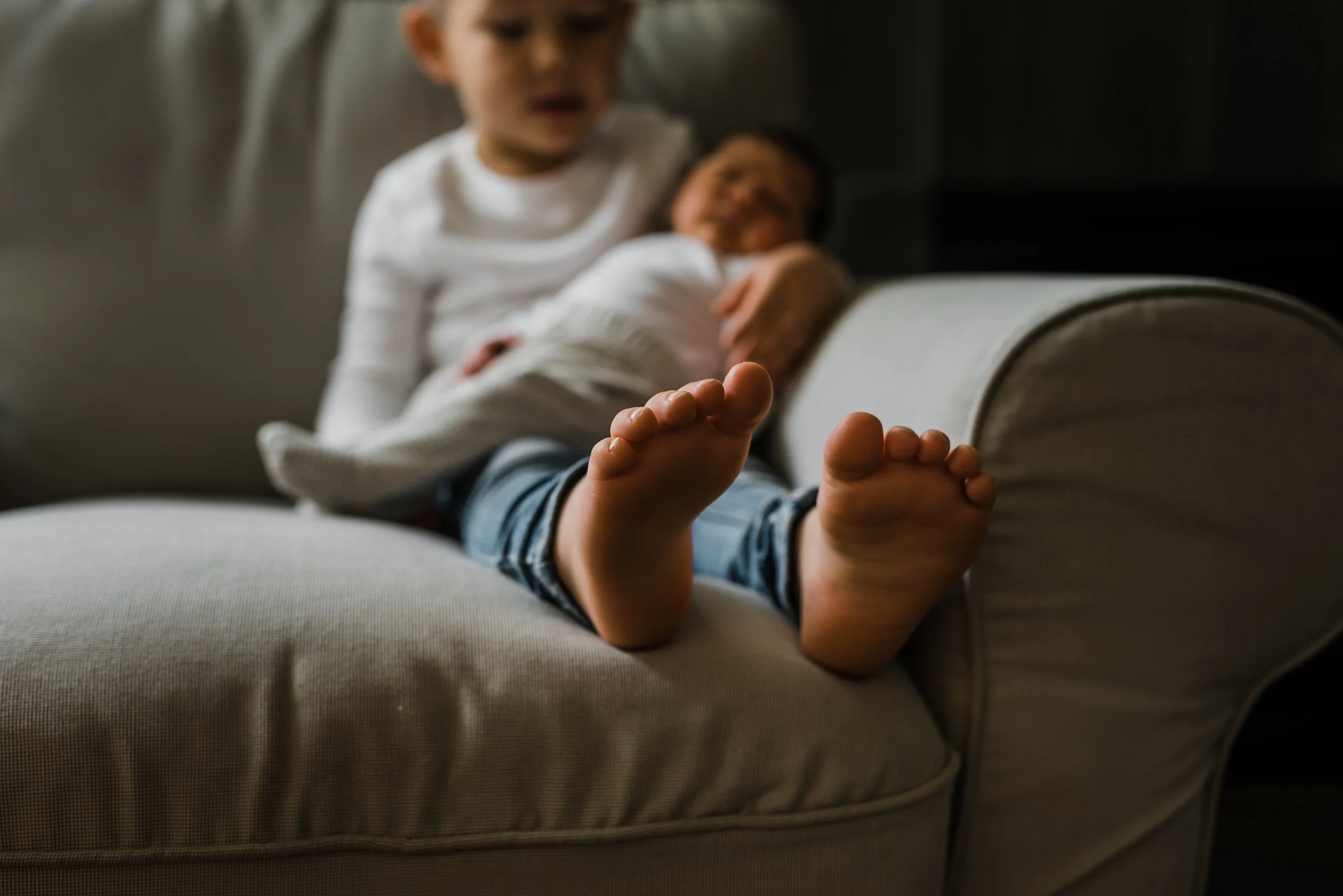 A young child and a woman, possibly her mother, are sitting on a beige couch in a living room. The child is in the foreground, wearing a white shirt and striped pants, and is reaching out with his foot towards the camera. The woman is in the background, smiling, wearing glasses and a white top. The room has dark walls and a cozy, relaxed atmosphere.