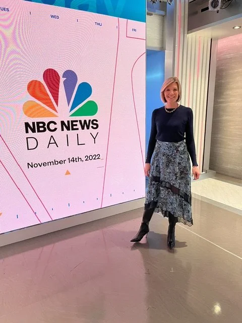 A woman is standing in front of a large NBC News Daily digital screen displaying the date November 14th, 2022. She is smiling and wearing a dark long-sleeve top, a patterned skirt, and high-heeled boots in an indoor setting.