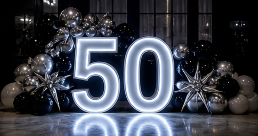 Illuminated 50 sign with black, white, and silver balloons and star-shaped balloon decorations on a dark backdrop.