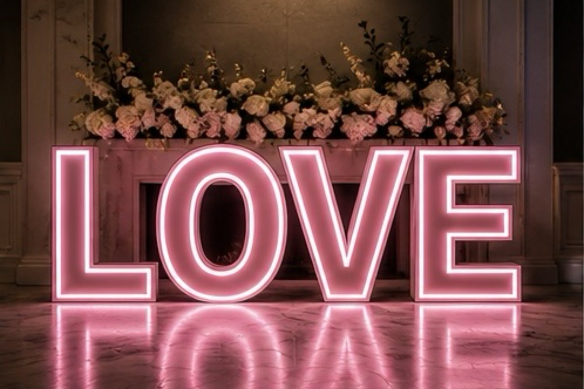 Neon pink sign spelling 'LOVE' in front of a pink floral arrangement on a fireplace mantel.