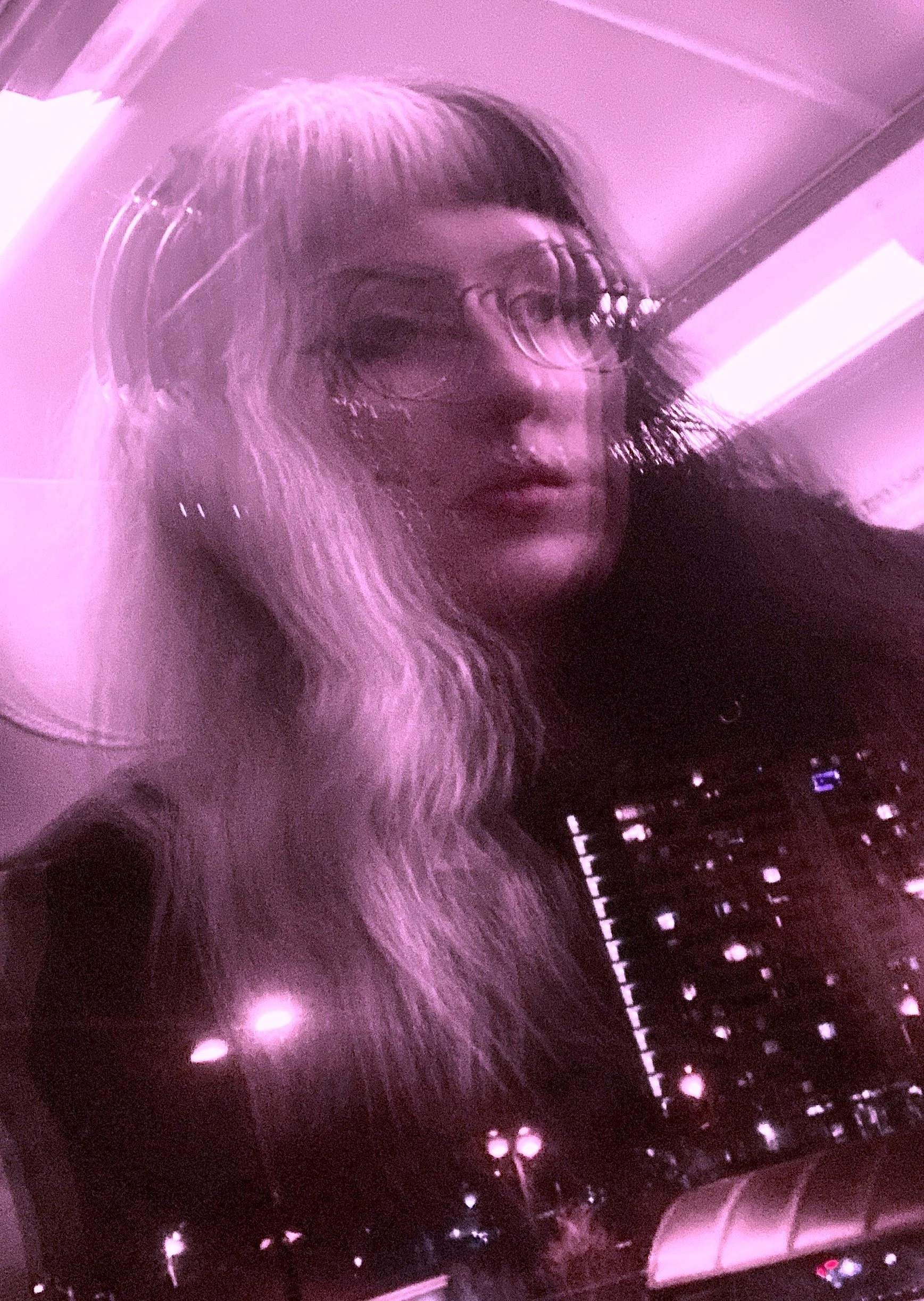 A nonbinary person with long hair and bangs taking a mirror selfie in a room with pink lighting, wearing glasses and a black top.