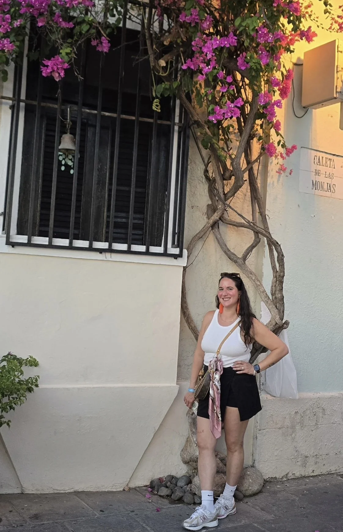 A woman standing next to a twisted tree with pink flowers, outside a building with a window and a sign that reads 'Caleta de las Monjas'. She is smiling, wearing a white tank top, black shorts, white sneakers, and has a scarf hanging from her bag.