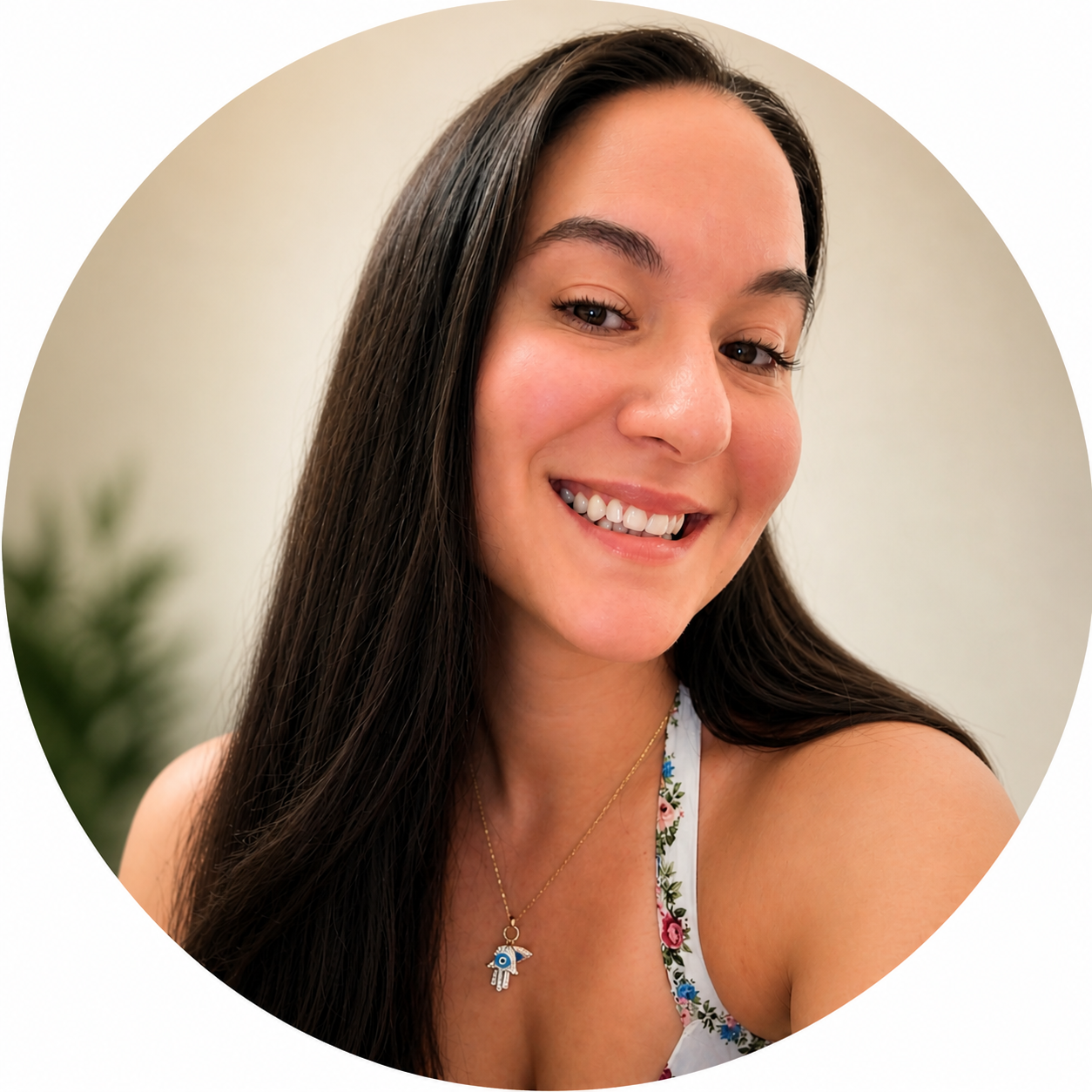 Close-up portrait of a smiling woman with long dark hair, wearing a floral dress and a necklace with a Hamsa hand pendant, against a neutral background.