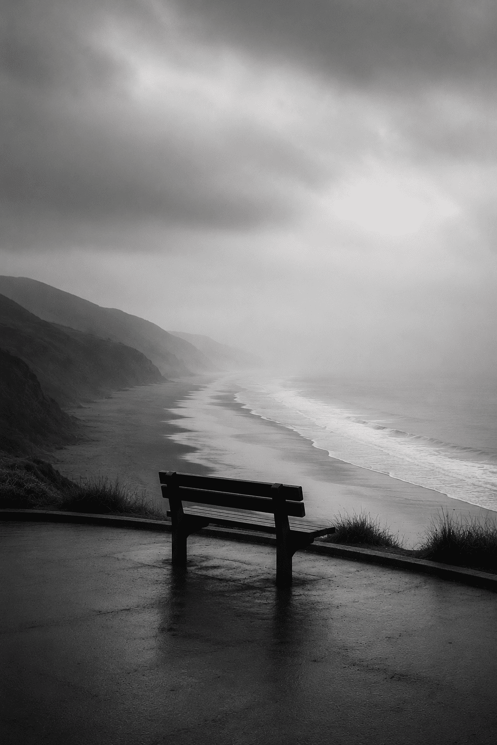Black and white photo of a solitary park bench overlooking a foggy, rocky coastline with waves crashing onto the shore under a cloudy sky.