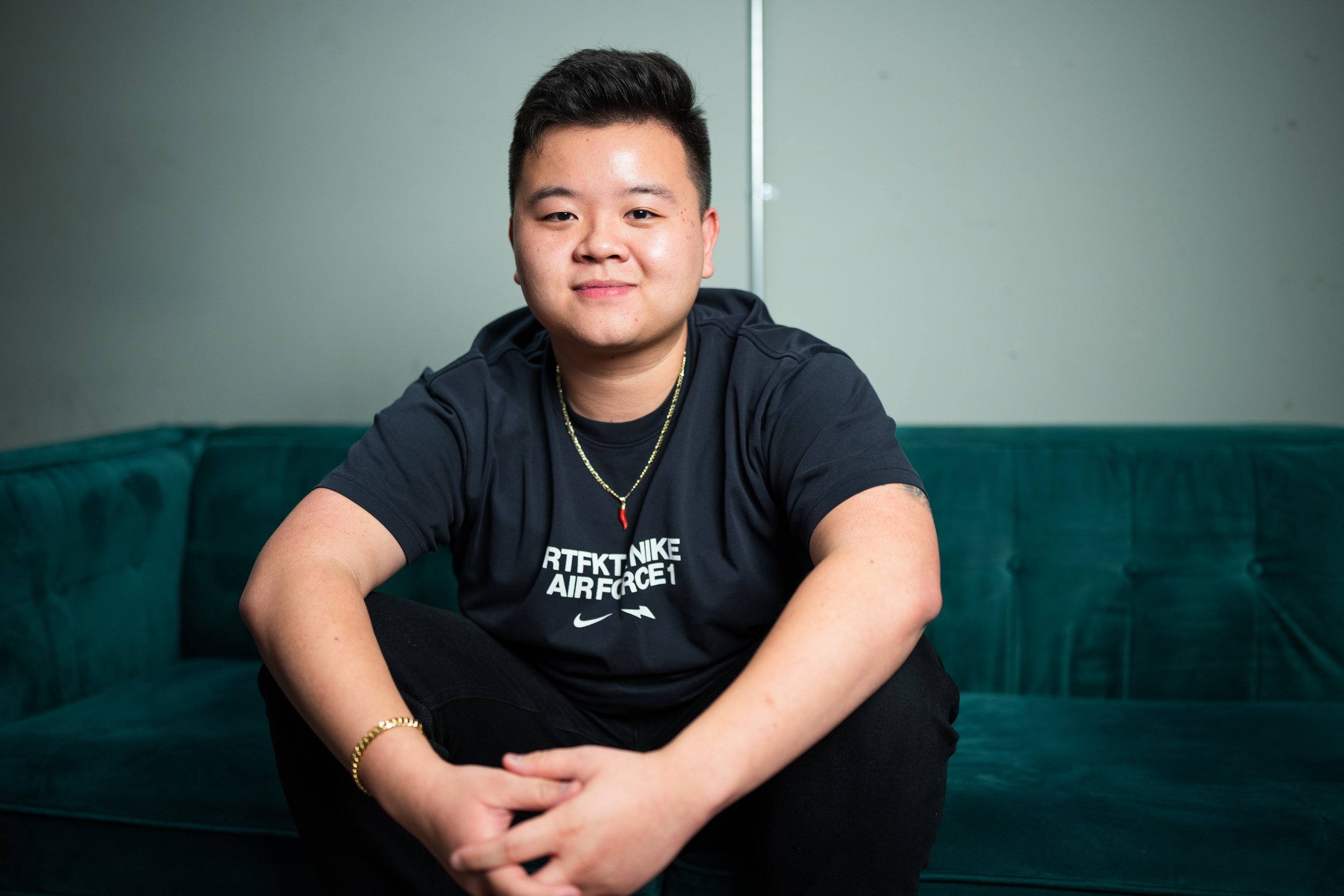 A young man sitting on a teal sofa in front of a mint green wall, wearing a black Nike shirt and smiling slightly at the camera.