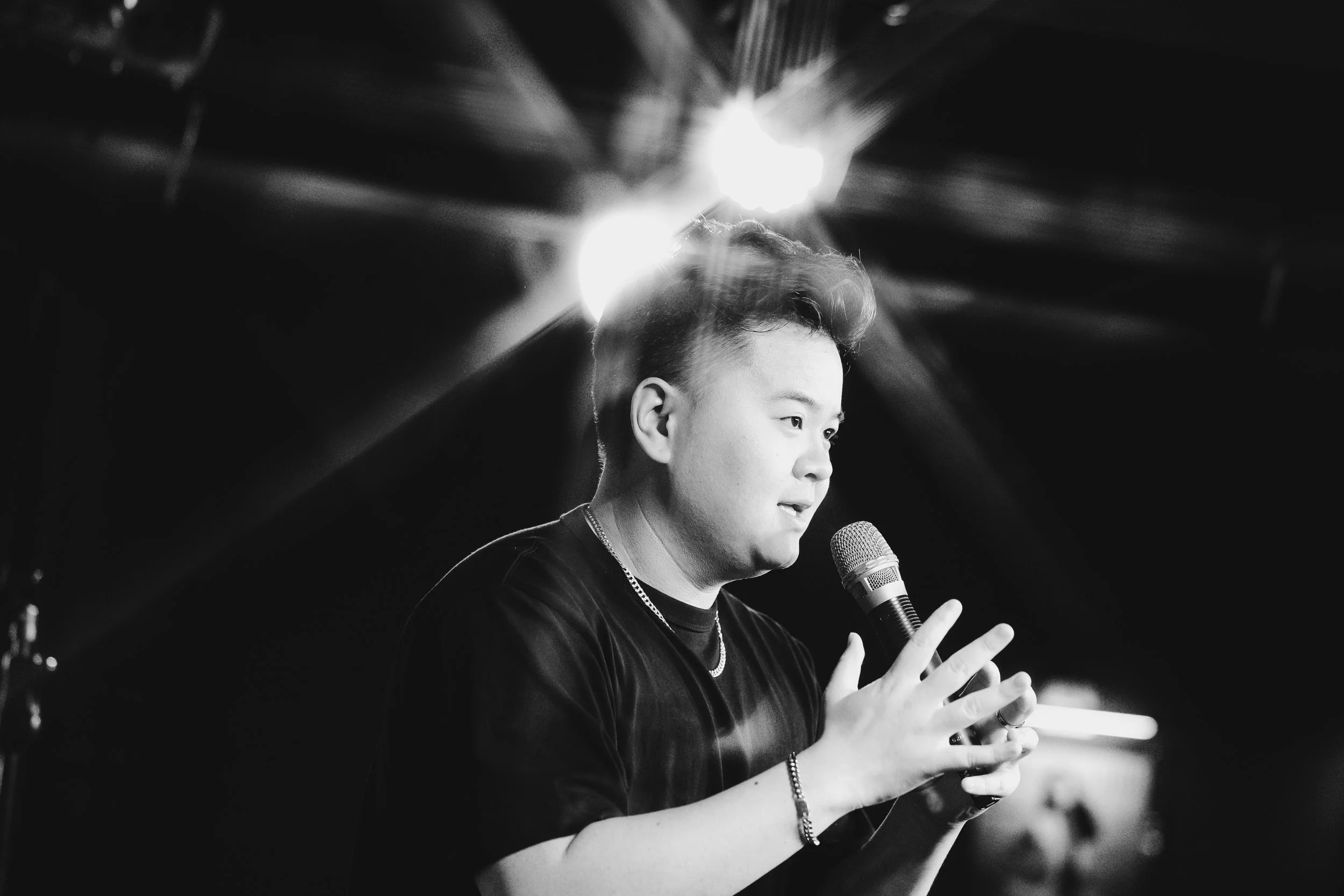A young man with short hair speaking into a microphone during a performance or speech, with bright lights shining in the background.