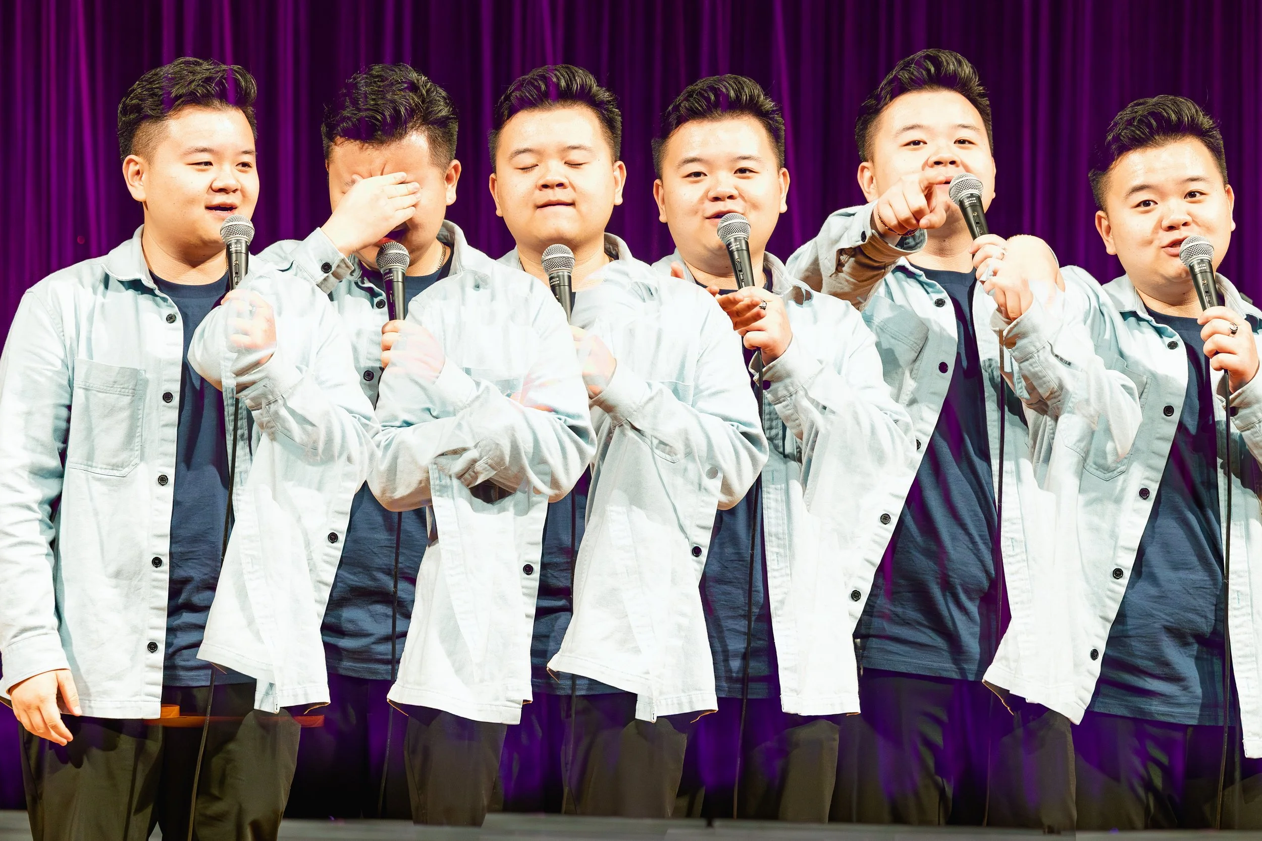 Seven young men standing on stage with purple curtain backdrop, holding microphones, dressed in matching light-colored jackets and dark shirts, engaging in a performance or speech.