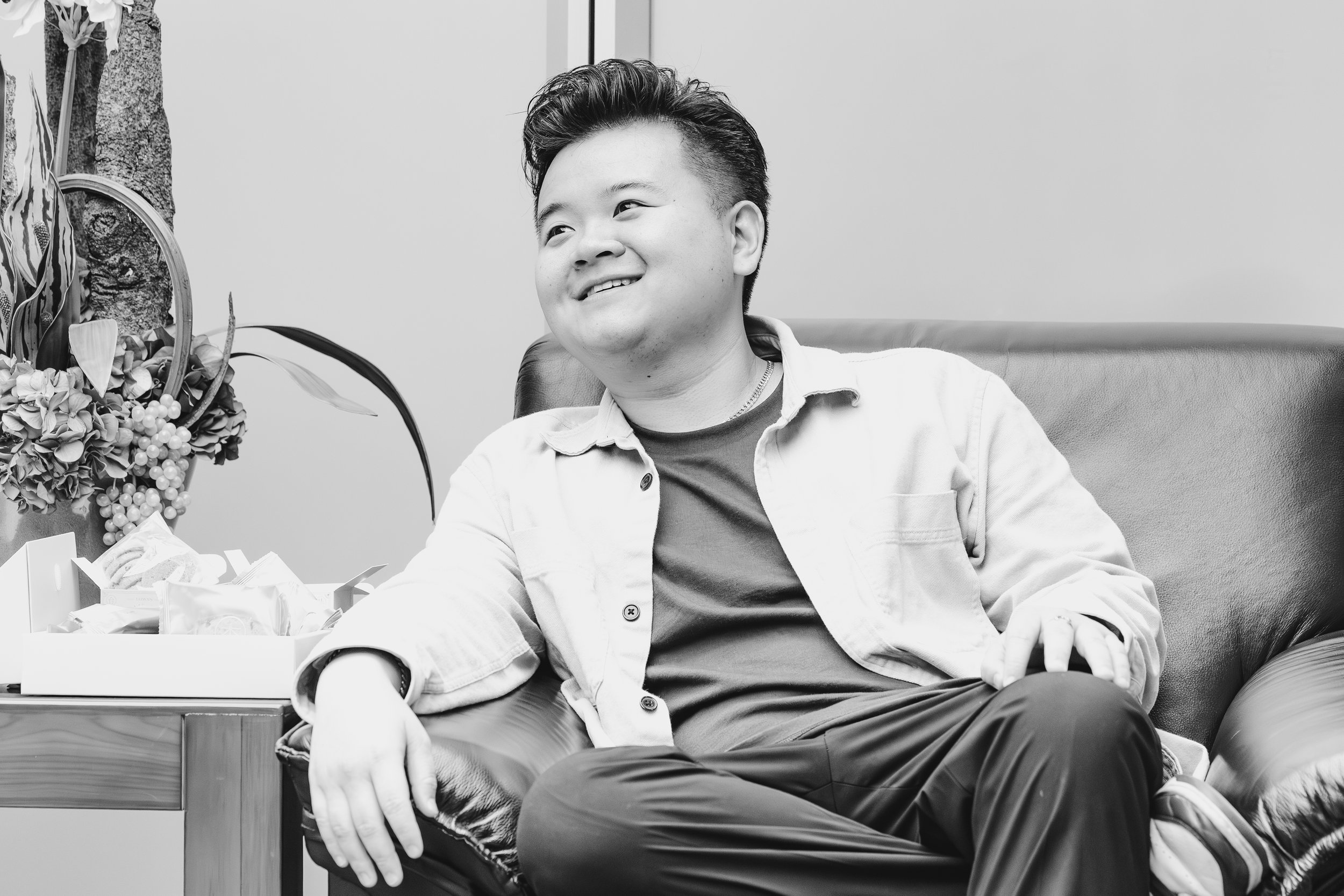 A young man with styled hair sitting on a couch, smiling, next to a table with a floral arrangement and various items, in black and white.