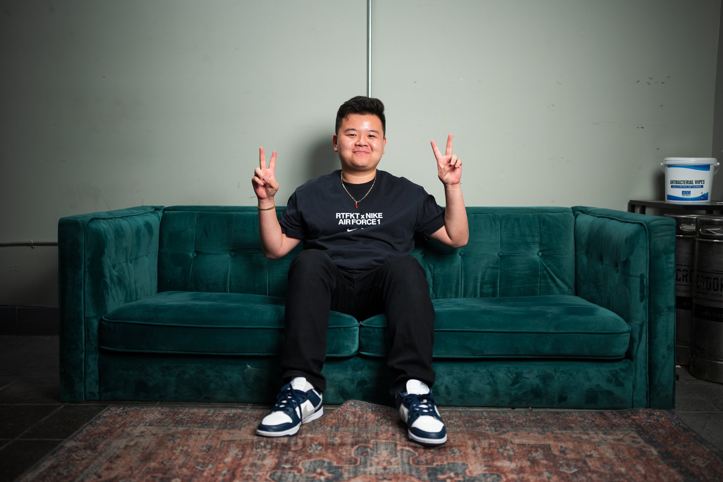 A young man sitting on a teal velvet sofa making peace signs with both hands, wearing a black Nike t-shirt, black pants, and sneakers, in a room with plain walls, a patterned rug, and a container of disinfectant wipes on a small table.